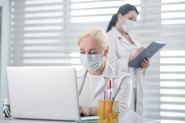 Médicos, colegas. Mulher loira, trabalhando em um laptop, morena com uma pasta perto da janela, em máscaras protetoras e jalecos brancos.