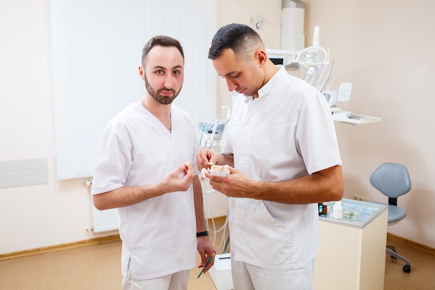 Los médicos dentistas están parados en la oficina, clínica con una silla, herramientas. Lugar de trabajo del dentista. Los ortodoncistas sostienen en sus manos y examinan el molde de la mandíbula. Concepto de odontología.