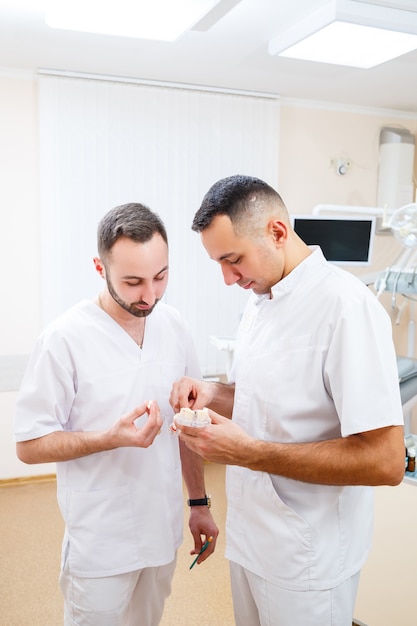 Los médicos dentistas están parados en la oficina, clínica con una silla, herramientas. Lugar de trabajo del dentista. Los ortodoncistas sostienen en sus manos y examinan el molde de la mandíbula. Concepto de odontología.