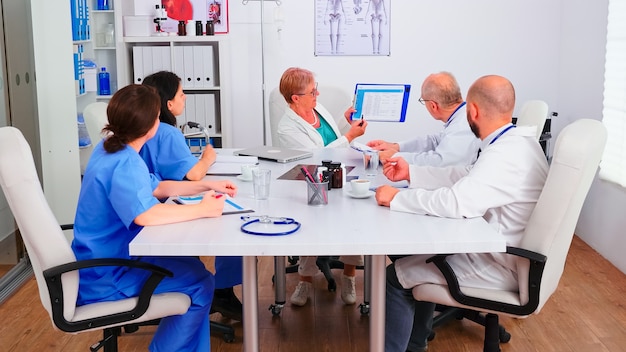 Médicos experimentados en la sala de reuniones del hospital trabajando juntos durante el seminario de salud, médico senior apuntando en el portapapeles. Terapeuta experto hablando con colegas sobre enfermedades, profesionales de la medicina