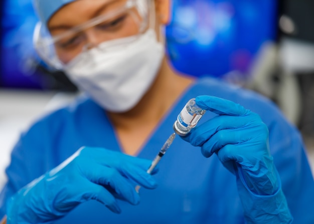 Médicos en ropa de uniforme azul con mascarilla de higiene facial sosteniendo la botella de la vacuna e insertando la aguja en ella, trabajando en el laboratorio.