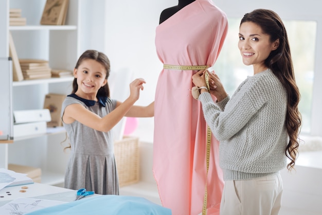 Medida da cintura. Mulher simpática e feliz segurando uma fita métrica e medindo a cintura enquanto desenha um vestido