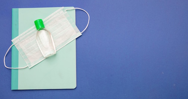 Foto medida de prevenção da propagação do coronavírus na escola máscara cirúrgica, gel de desinfecção com álcool e caderno em fundo azul