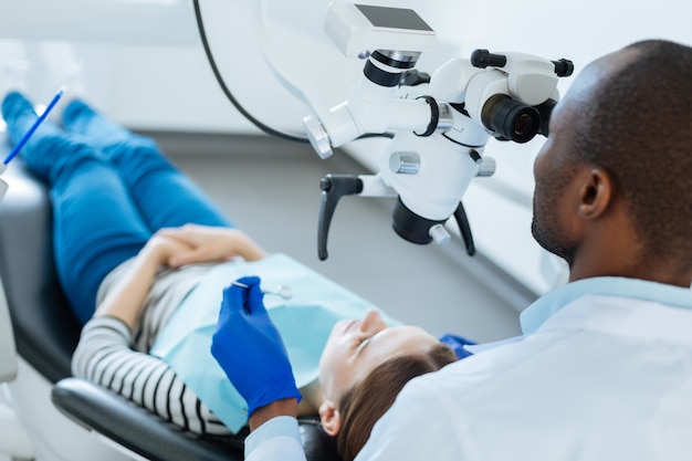Medida necessária. Dentista simpática e habilidosa verificando os dentes de seus pacientes em busca de microfissuras com a ajuda de um microscópio enquanto ela está deitada na cadeira do dentista