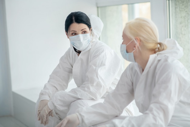 Medidas de protección. Dos trabajadoras médicas con ropa protectora y máscara médica sentada junto a la ventana, discutiendo algo