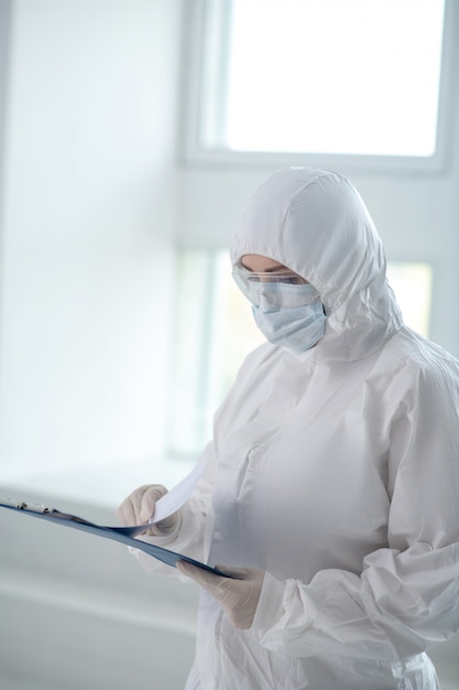 Medidas de protección. Trabajador médico en ropa protectora, máscara médica y gafas mirando a través de documentos en la carpeta de clip