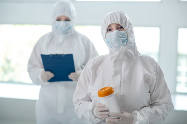 Foto medidas de protección. trabajador médico en ropa protectora y máscara médica sosteniendo la botella de toallitas desinfectantes, su colega de pie con una carpeta de clip