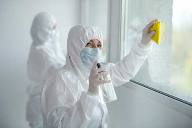 Medidas protetoras. Trabalhadores médicos em roupas de proteção e máscaras médicas, limpeza e desinfecção de vidraças