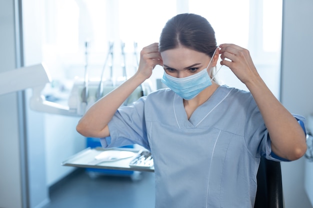 Medidas de seguridad. Encantadora dentista alegre de pie en su oficina y poniéndose una mascarilla mientras se prepara para una cita con un paciente