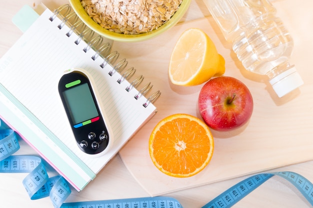 Medidor de glucosa en un bloc de notas, tazón de avena, cinta métrica, frutas, una botella de agua en una mesa de madera