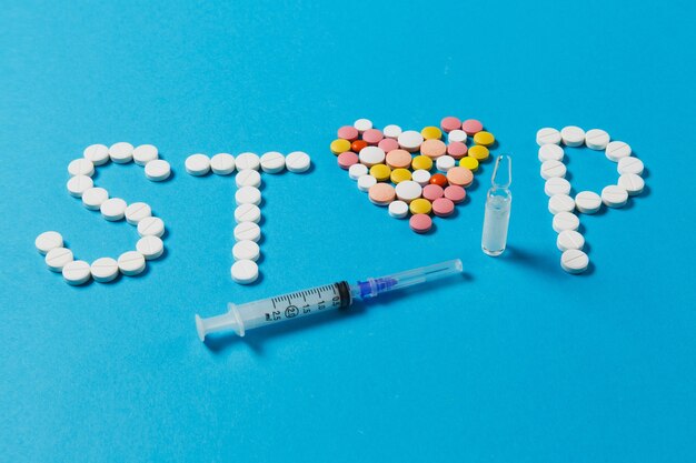 Medikamente weiße, bunte runde Tabletten in Wort Stop auf blauem Hintergrund isoliert. Pillenherz, Ampulle, leere Spritzennadel. Konzept der Behandlungswahl, gesunder Lebensstil. Platzwerbung kopieren.