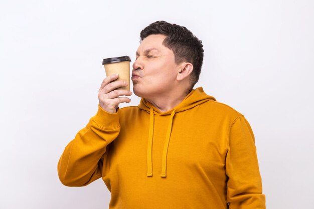 Medio alegre con cabello oscuro besando una taza desechable de café para llevar disfrutando de una bebida