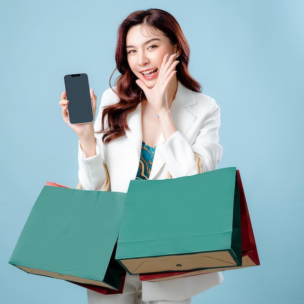 Medio cuerpo de una hermosa mujer de negocios asiática con traje blanco con la boca abierta levantando las manos gritando un anuncio que muestra una aplicación de teléfono móvil y una bolsa de compras en un fondo azul aislado