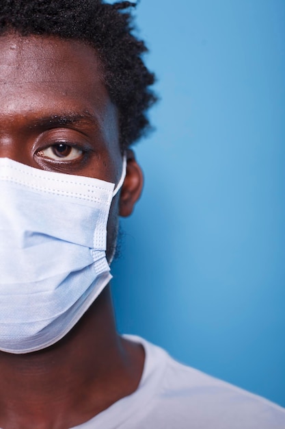 Medio rostro de primer plano de un hombre negro con máscara mirando fijamente a la cámara contra un fondo azul Imagen recortada de un individuo afroamericano que usa una máscara facial para protegerse del coronavirus