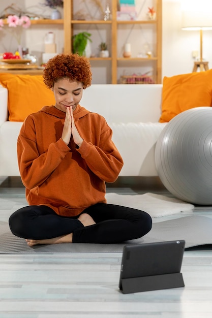 Meditação de atenção plena de ioga Jovem garota africana saudável praticando ioga em casa Mulher sentada em pose de lótus no tapete de ioga olhos fecham as mãos em oração meditando interior Menina fazendo prática de respiração