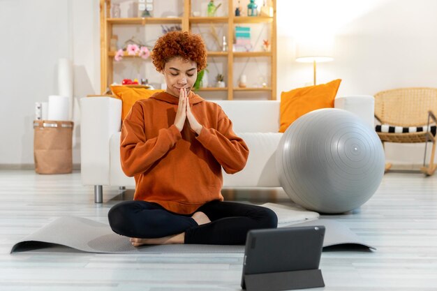 Meditación de atención plena de yoga Joven niña africana saludable practicando yoga en casa Mujer sentada en posición de loto en la estera de yoga ojos cerrar las manos en oración meditando interior Niña haciendo práctica de respiración