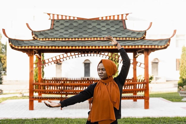 Meditación china Qigong y entrenamiento deportivo al aire libre. La mujer musulmana negra está meditando al aire libre cerca del cenador chino.