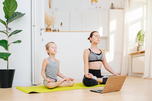Meditando a jovem mãe e seu filho fazendo ioga juntos sentados em lótus asana com os olhos fechados em uma cozinha espaçosa