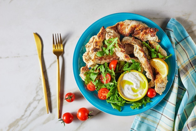 Mediterrane gegrillte Hähnchenstreifen serviert mit gebackenen weißen Kartoffeln und Salat aus Rucola und Kirschtomaten auf blauem Teller mit Besteck und Tzatziki-Sauce auf heller Marmoroberfläche, Draufsicht