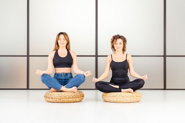 Meditierende Frauen, die im Lotus sitzen, posieren mit geschlossenen Augen im weißen Raum. Es ist Yoga-Zeit.