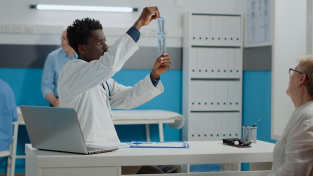 Mediziner afrikanisch-amerikanischer Abstammung analysiert Röntgenaufnahme der Patientenversorgung am Schreibtisch sitzend. Schwarzer Arzt, der einen Röntgenscan für professionelle Diagnosen und Ergebnisse im medizinischen Kabinett hält