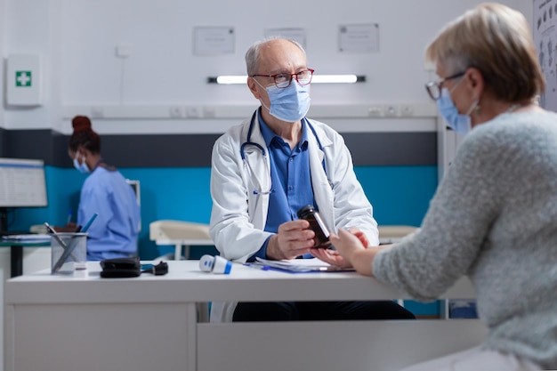 Mediziner, der eine Flasche Pillen für eine ältere Frau mit Gesichtsmaske bei einem Arztbesuch hält. Arzt bei der Untersuchung, der dem Patienten Medikamente im Glas zur Behandlung während einer Coronavirus-Pandemie zeigt.