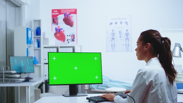 Foto mediziner schreiben diagnose auf computer mit greenscreen-monitor, während die krankenschwester im hintergrund notizen macht. computer mit austauschbarem bildschirm, der von medizinspezialisten im krankenhaus verwendet wird und uniform trägt