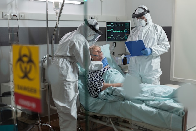 Medizinische Krankenschwester in einem PSA-Anzug, die einem älteren Patienten im Laufe der globalen Pand...