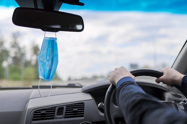 Medizinische Schutzmaske des Fahrers im Auto, die Hände des Mannes am Lenkrad des fahrenden Fahrzeugs