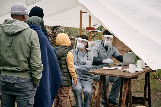 Medizinische Spezialisten für Schutzmasken, die am Tisch im Zelt sitzen und den Gesundheitszustand von Migranten überprüfen, die in der Schlange stehen