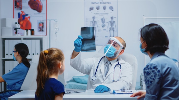 Medizinischer Mitarbeiter mit Schutzmaske, der die Röntgenaufnahme des Elternteils während des Coronavirus erklärt. Facharzt für Medizin, der Beratung im Gesundheitswesen anbietet, Röntgenbehandlung im Klinikkabinettkrankenhaus