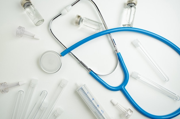 Medizinisches Layout auf weißem Tisch, flach gelegen. Blaues Stethoskop oder Phonendoskop, umgeben von Fläschchen, Ampullen und Reagenzgläsern.