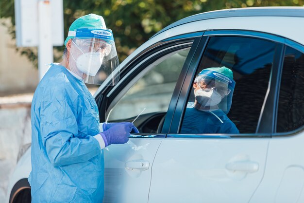 Medizinisches Personal, das eine PSA trägt und eine PCR mit einem Tupfer in der Hand an einem Patienten in seinem Auto durchführt, um festzustellen, ob er mit COVID-19 infiziert ist