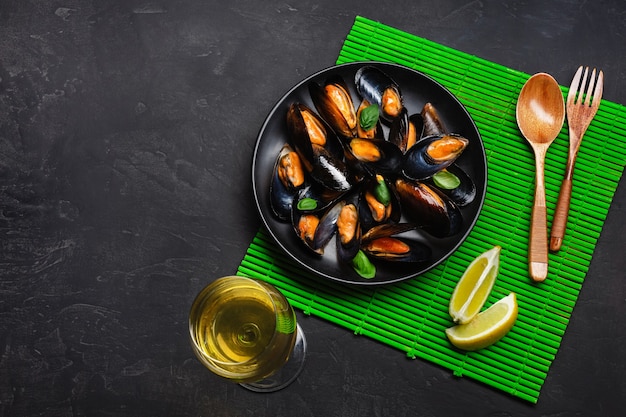 Meeresfrüchtemuscheln, Basilikumblätter in einem schwarzen Teller mit Weinglas, Zitrone, Holzlöffel und Gabel auf grüner Bambusmatte und Steintisch. Ansicht von oben.