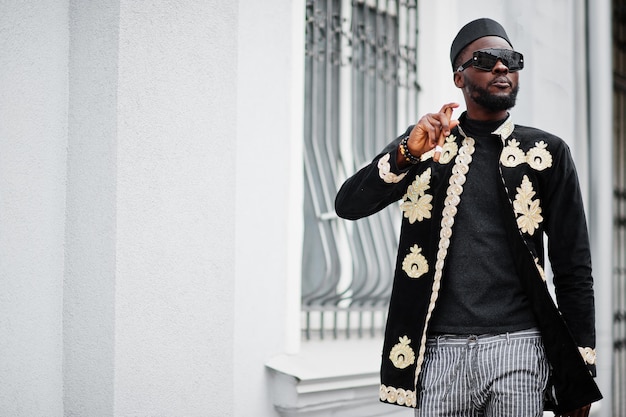 Mega stylischer afrikanischer Mann in traditioneller Jackenhaltung. Modischer schwarzer Typ mit Hut und Sonnenbrille mit Zigarre in der Hand.