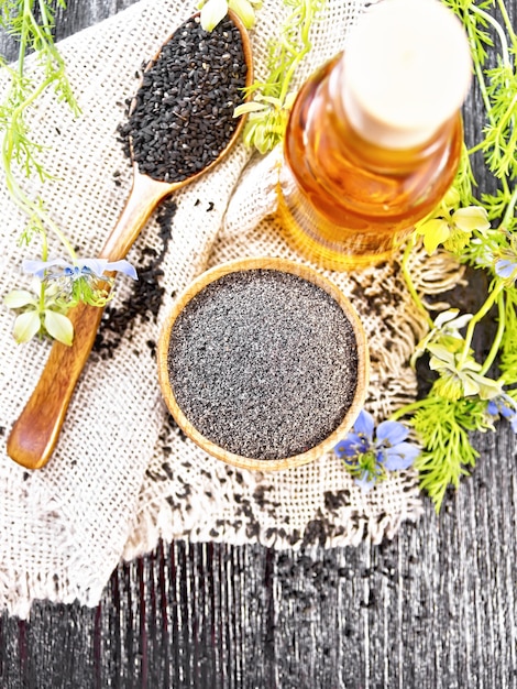 Mehl aus schwarzem Kümmel in einer Schüssel, Samen in einem Löffel auf Sackleinen, Öl in der Flasche und Zweige Nigella sativa mit blauen Blüten und grünen Blättern auf dunklem Holzbretthintergrund von oben