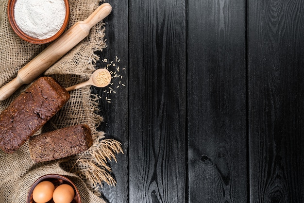 Mehl in einer Holzschale auf dunklem Holztisch mit Ährchen von Weizen, Eiern und Milch, Draufsicht mit Kopierraum