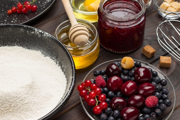 Mehl und Beeren. Glas Honig und Marmelade. Zutaten zum Backen von Beerenkuchen. Dunkle Holzoberfläche. Draufsicht