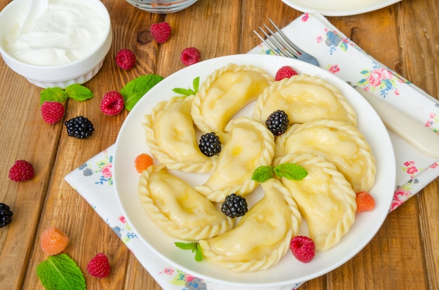 Mehlklöße mit Hüttenkäse, frischen Beeren und Sauerrahm auf einer weißen Platte. Traditionelles ukrainisches und russisches Gericht.