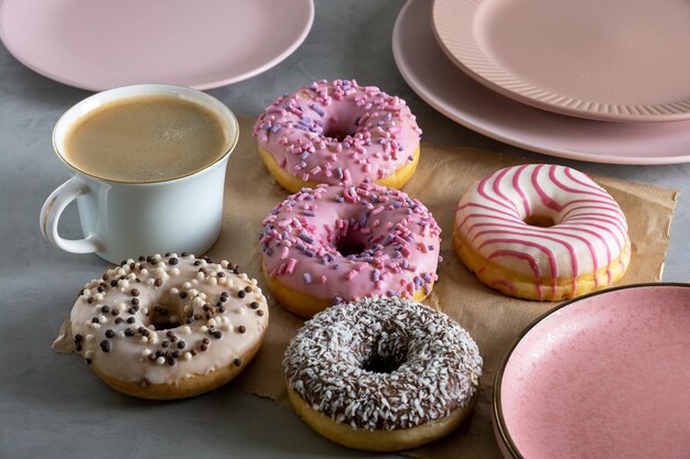 Mehrere Donuts liegen auf Pergamentpapier auf grauer Oberfläche Porzellantasse Kaffee und rosafarbene Teller stehen neben Donuts, die auf dem Tisch ausgelegt sind Selektiver Fokus