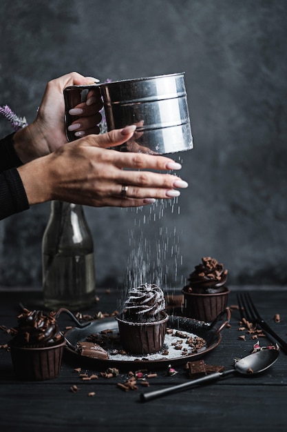 Mehrere Muffins oder Cupcakes mit schokoladenförmiger Sahne am schwarzen Tisch. Die Hand einer Frau zerbröckelt Puderzucker auf einem Kuchen.