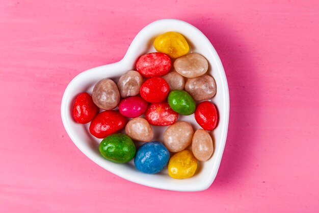 Mehrfarbige Bonbons in einem Teller in Form eines Herzens auf rosafarbenem Holzhintergrund. Valentinstag.