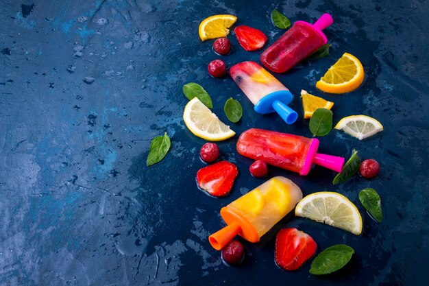 Mehrfarbiges helles Frucht-Eis am Stiel mit Erdbeere, Kirsche, Zitrone, Orange, Zitrone und Minze und schneidet frische Früchte auf einem dunklen Blau