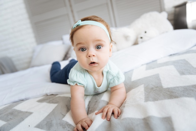 Mein Zimmer erkunden. Nettes positives kleines Mädchen, das auf dem Bett im Schlafzimmer liegt und auf das Publikum schaut, während Interesse ausdrückt