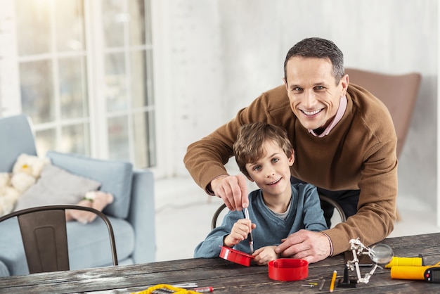 Meine Unterstützung. Hübscher fröhlicher kleiner blonder Junge, der lächelt und lernt, wie man Instrumente benutzt, und sein Vater, der hinter ihm steht und ihm hilft