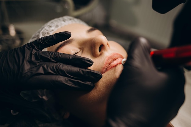 Meister im Schönheitssalon malt Lippen von Mädchenmodell mit permanenter Make-up-Maschine Nahaufnahmeansicht