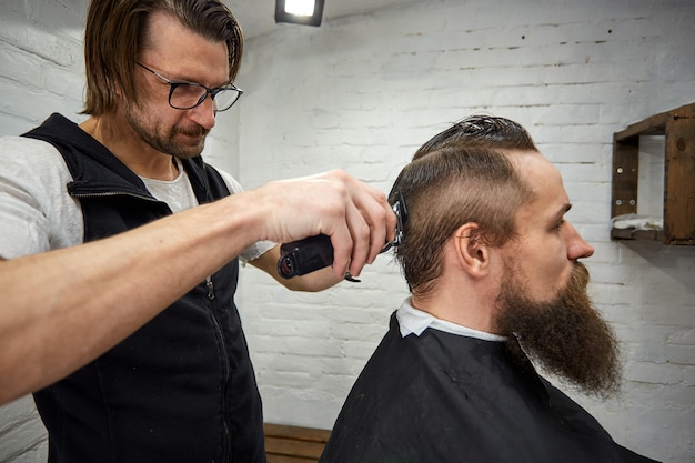 Meister schneidet Haare und Bart von Männern im Friseursalon, Friseur macht Frisur für einen jungen Mann