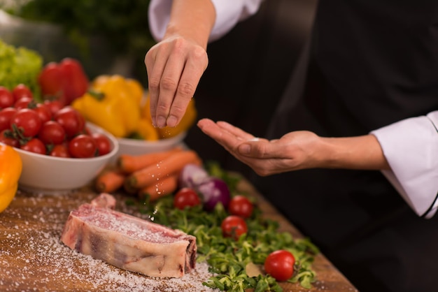 Meisterkoch übergibt Salz auf ein saftiges Stück rohes Steak mit Gemüse auf einem Holztisch