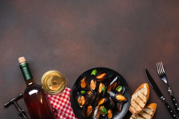 Mejillones de marisco y hojas de albahaca en un plato negro con botella de vino, copa de vino, sacacorchos, tenedor y cuchillo sobre una toalla y fondo oxidado. Vista superior con lugar para su texto.