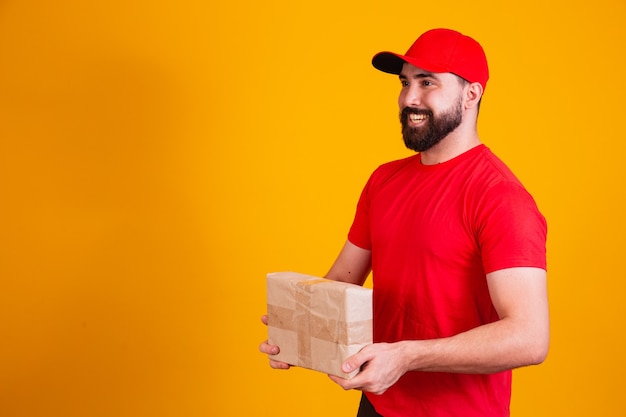 Mejor concepto de servicio. Repartidor caucásico sonriente en gorra roja y apilamiento sosteniendo la pila de cajas de cartón mientras está de pie aislado sobre fondo amarillo de estudio. Paquetes de transporte de correo